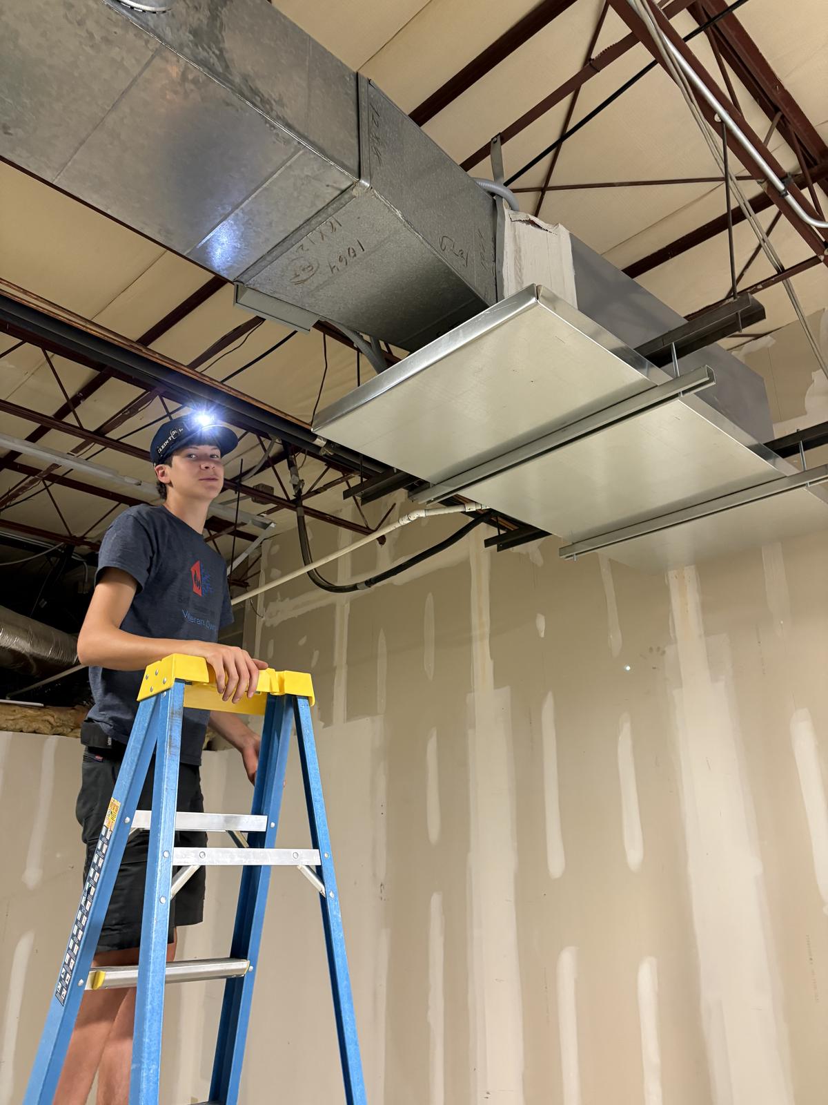 Sid's Heating & Air technician on a ladder servicing a unit in a light commercial building near Monroe NC